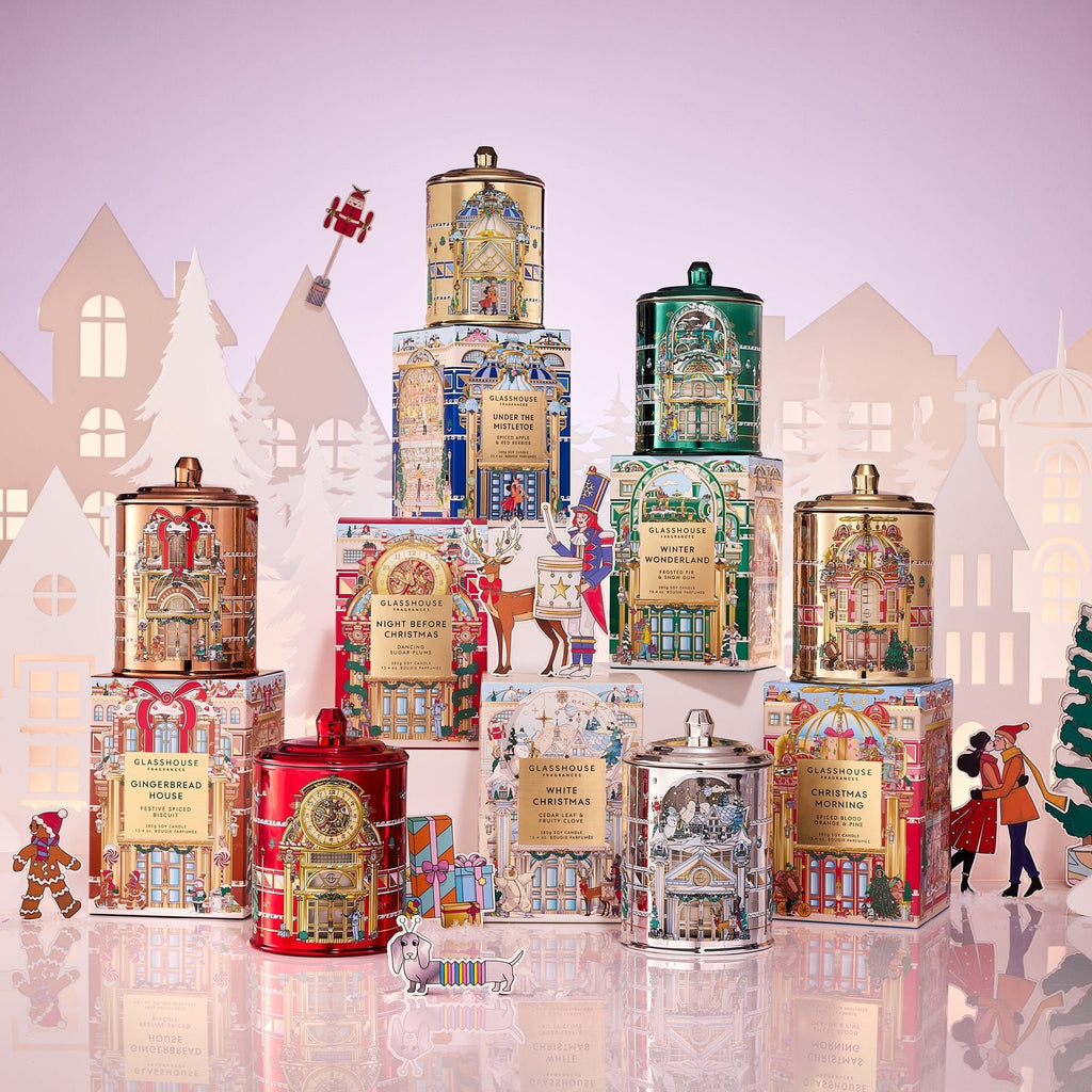 Glasshouse Fragrances’ Christmas Morning 13.4oz Candle is displayed with vibrant holiday tea tins, festive boxes, paper houses, a snowman and figures, creating a joyful winter scene.