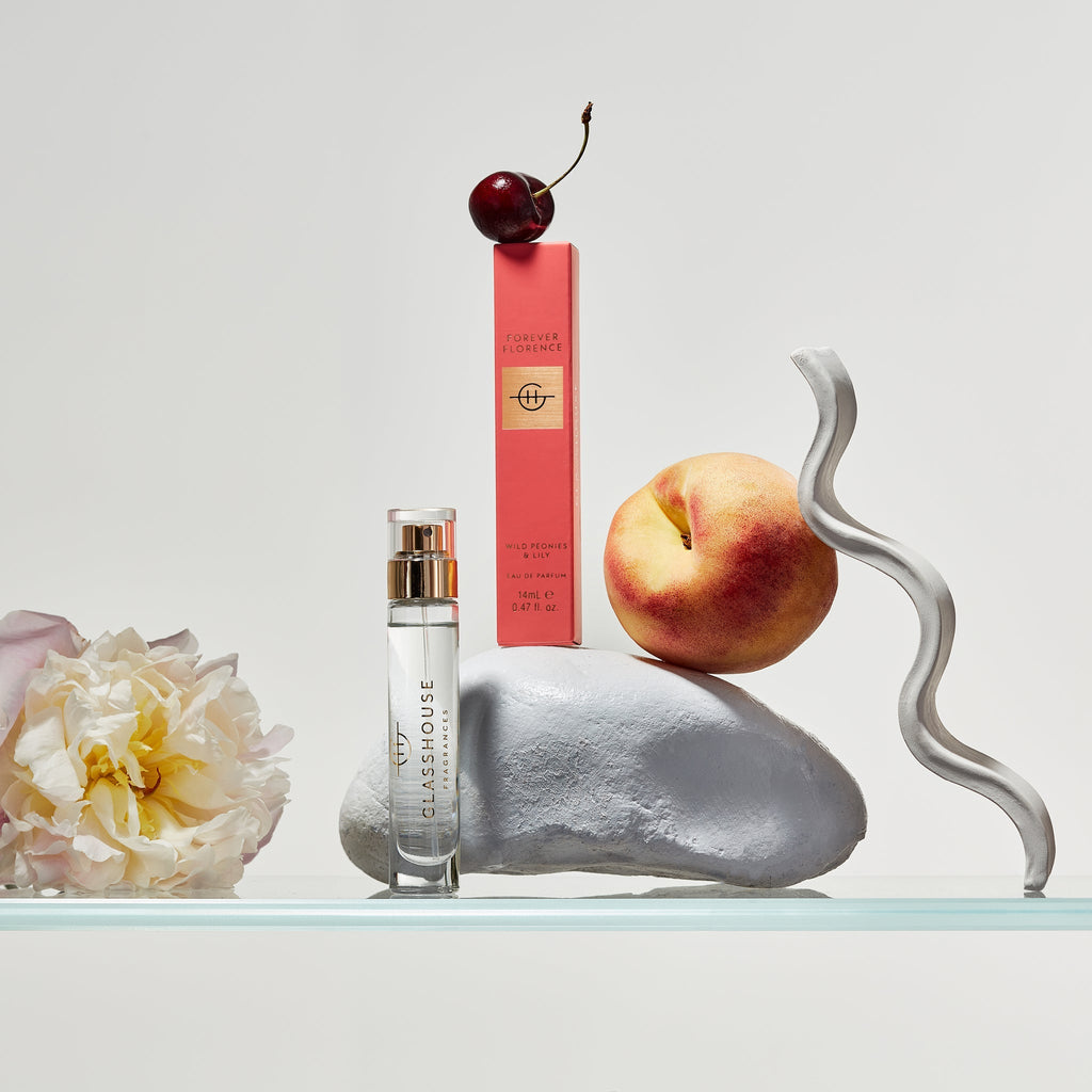 The Glasshouse Fragrances Eau de Parfum 14ml - Forever Florence bottle and packaging rest elegantly on a glass surface, accented by a peach, pink peonies, a curved metal strip, and a cherry on a rock. This minimalist scene captures the sophisticated essence of the exquisite fragrance against a light background.