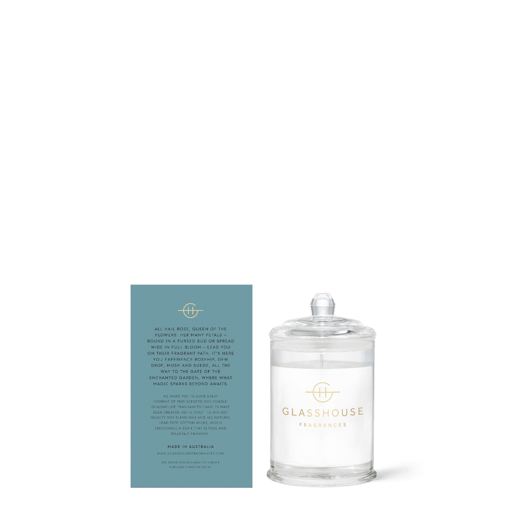 A clear glass candle labeled Glasshouse Fragrances is next to a teal card on a white background. This 2.21oz triple scented soy candle, named Enchanted Garden by Glasshouse Fragrances, entices you with scents of climbing and rambling roses.