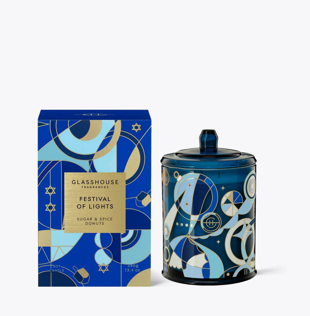 The Glasshouse Fragrances - Festival of Lights 13.4oz Candle sits beside its box, both adorned with festive blue and gold geometric patterns. Made from a non-toxic soy blend wax, it features the Sugar & Spice Donuts scent. The candles glass container mirrors the boxs design.