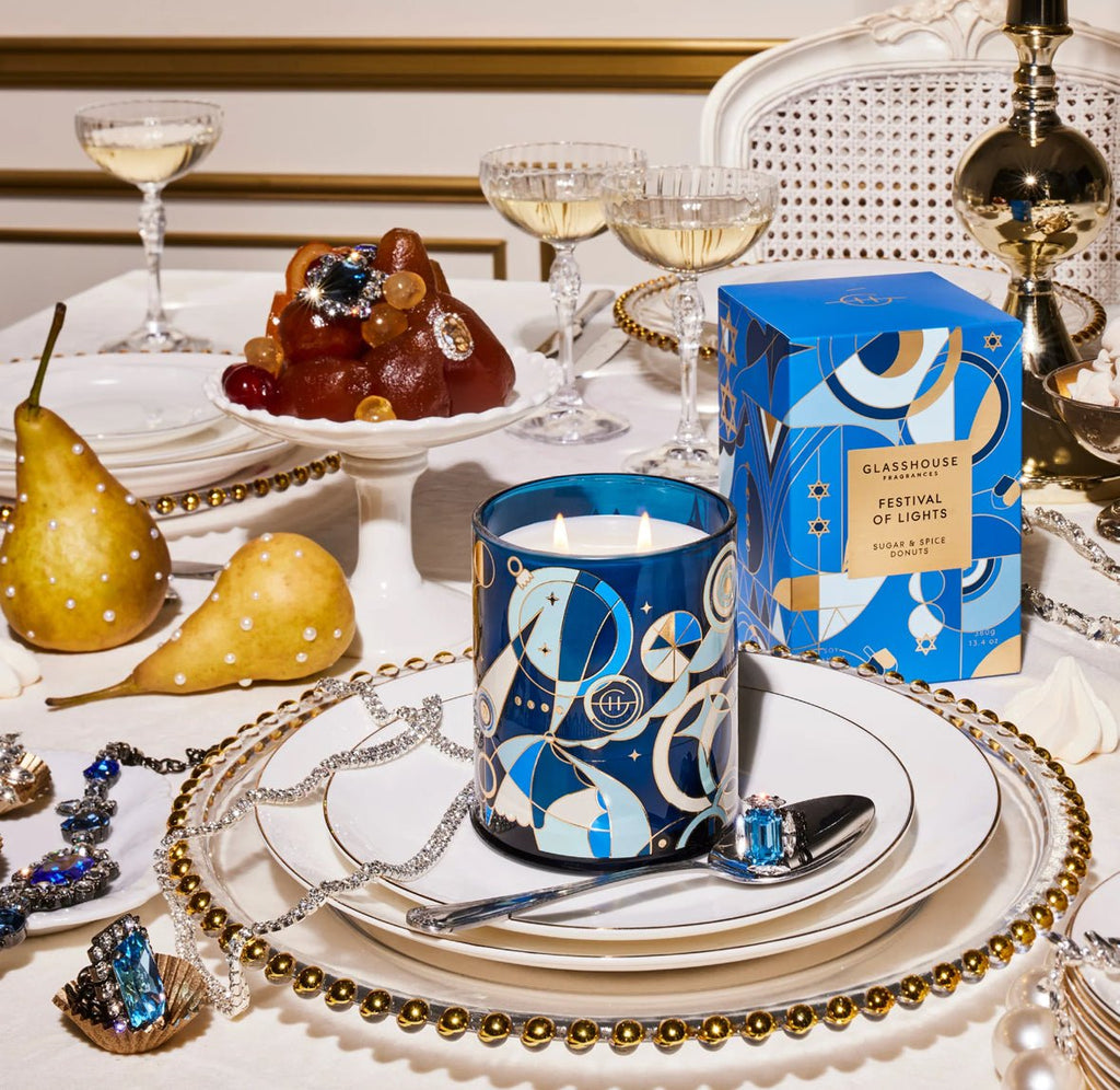 A festive table features Glasshouse Fragrances Festival of Lights 13.4oz Candle with a blue and gold design, complemented by non-toxic soy blend wax pears, a bejeweled dessert, and beaded strands on white plates.