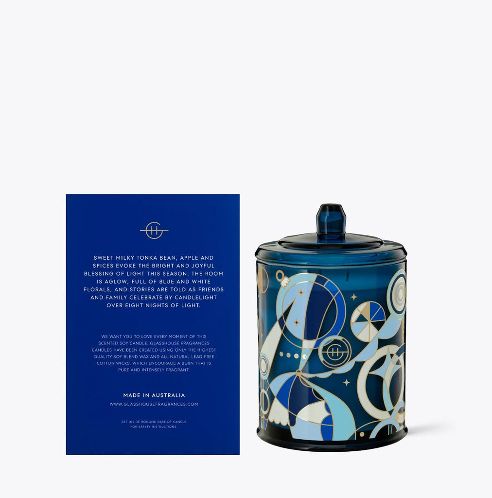 The Glasshouse Fragrances - Festival of Lights 13.4oz Candle features a candle jar with gold and blue abstract circles on non-toxic soy blend wax. It comes in a blue box with white text detailing the Triple Scented Candles aroma and origin, set against a plain background.