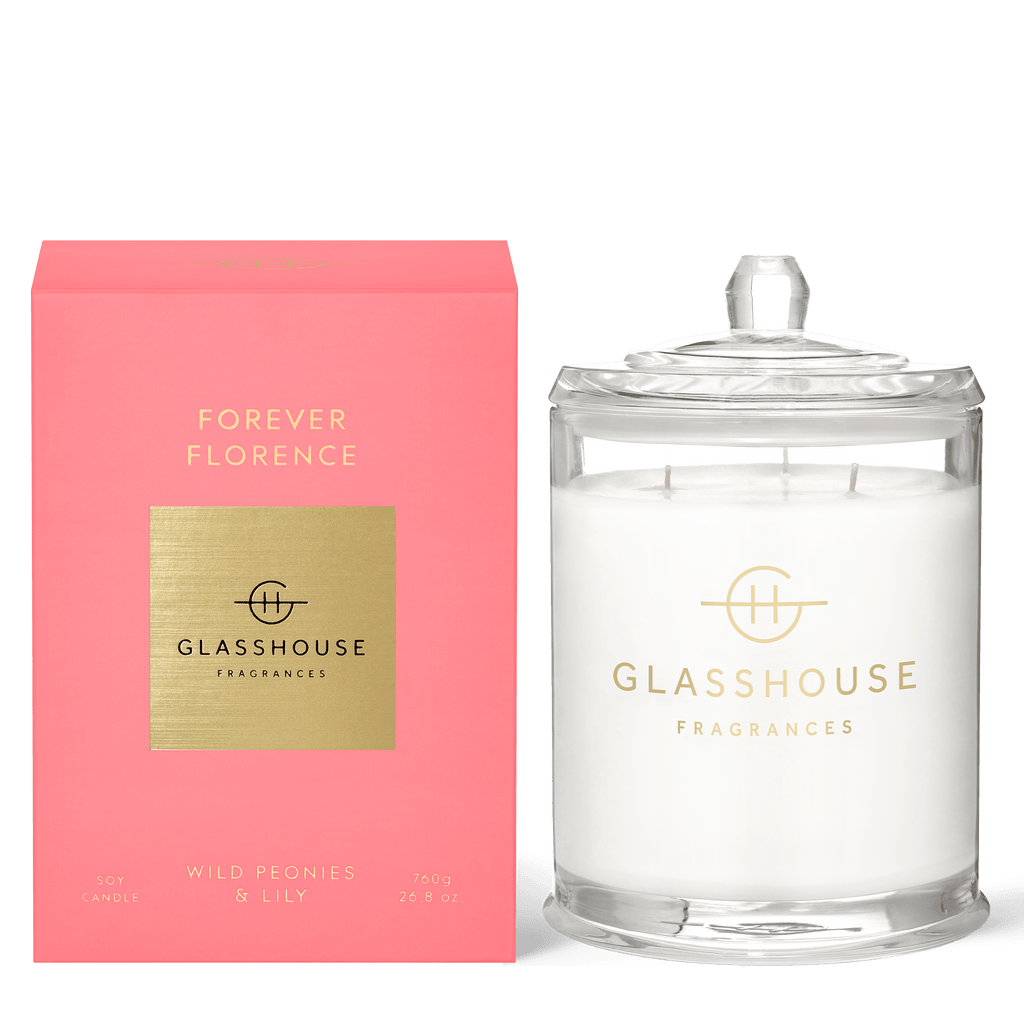 A clear glass jar soy candle with a lid, labeled with gold branding, stands next to a pink box featuring Forever Florence and Glasshouse Fragrances. The white candle is infused with peony and lily scents.