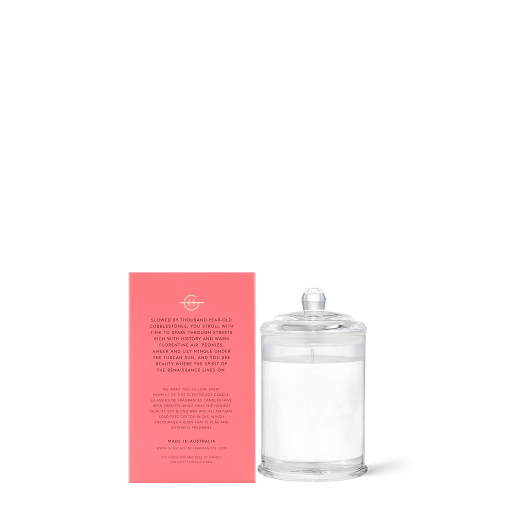 The Glasshouse Fragrances - Forever Florence Mini 2.1oz Candle in a clear jar is set next to its pink rectangular box with white text, both infused with jasmine and peony notes, elegantly displayed against a plain white background.