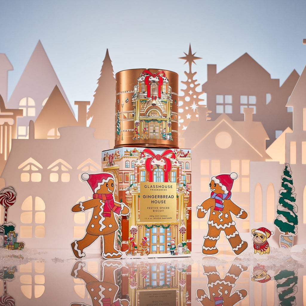 Two Glasshouse Fragrances Gingerbread House 13.4oz candles are stacked among paper house cutouts and festive gingerbread figures, set on snowy ground with a wintry backdrop—perfect for a holiday candle display.