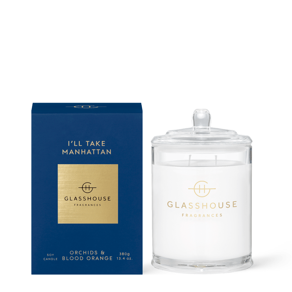 The Glasshouse Fragrances Ill Take Manhattan candle, in a blue and gold box, stands beside a clear glass jar with a lid. This 13.4oz orchid and blood orange candle features a delightful soy blend wax that enhances its enchanting aroma.