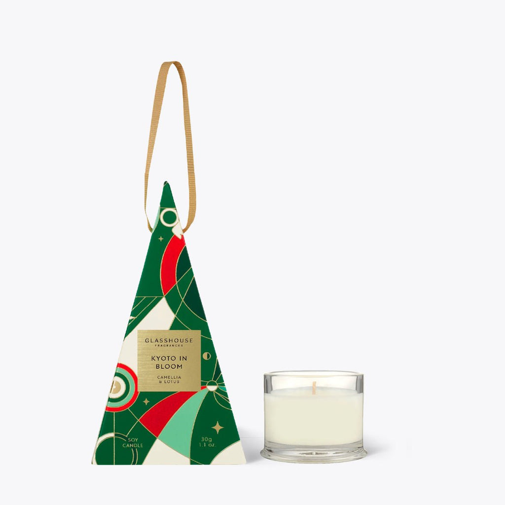 A small glass candle bauble labeled Kyoto in Bloom, Glasshouse Fragrances sits beside a triangular green, red, and cream package adorned with floral and geometric designs. The brown-ribbon-topped packaging captures the essence of Camellia & Lotus.