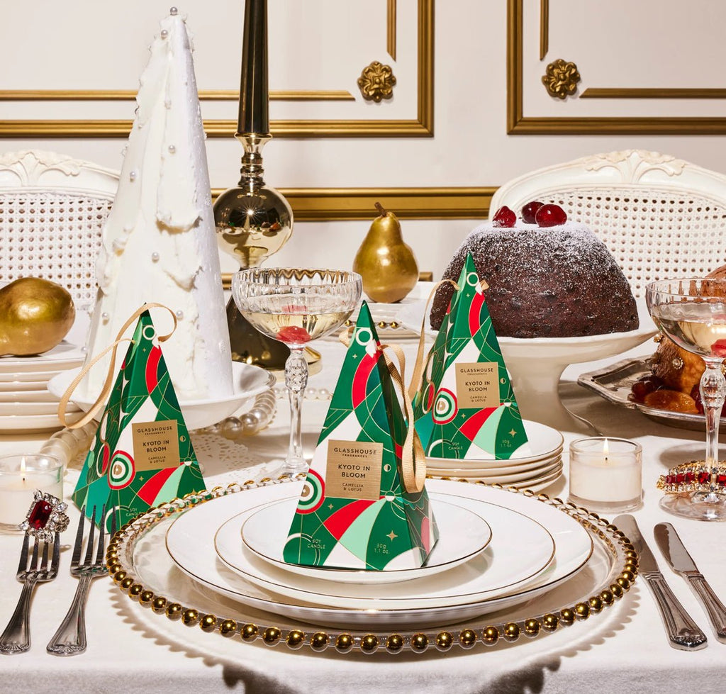 A festive dining table features white and gold plates, silverware, and crystal glasses. Green and red pyramid-shaped gift boxes rest on plates alongside a Glasshouse Fragrances Kyoto in Bloom Bauble. A large white tree and cake adorn the background, set against an elegant gold-trimmed wall.