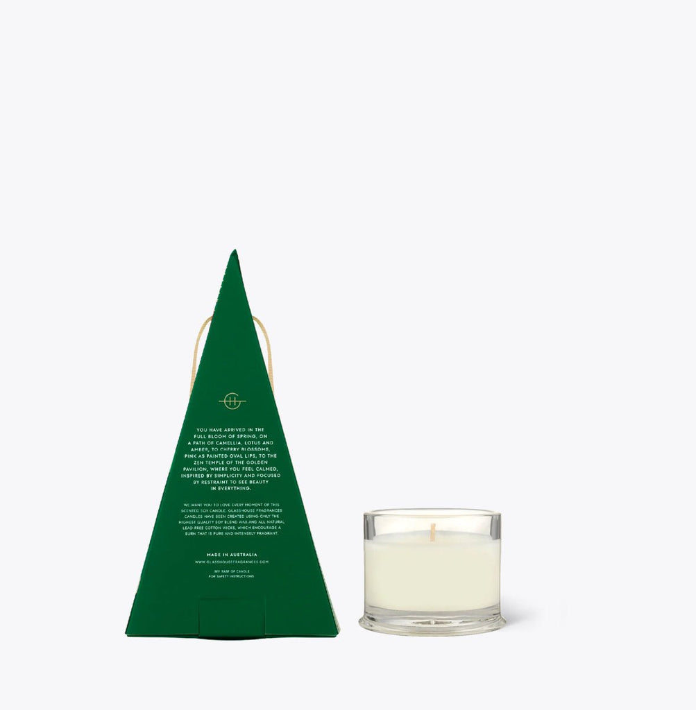 Next to a small, clear glass Kyoto in Bloom candle with white wax is a green pyramid package with white text and a handle. The Glasshouse Fragrances - Kyoto in Bloom Bauble elegantly enhances any space.