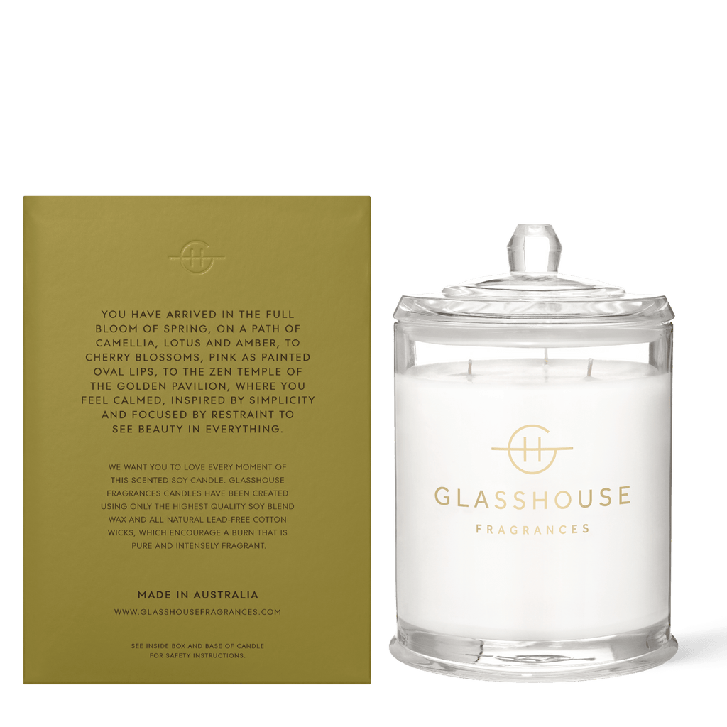 The Glasshouse Fragrances Kyoto in Bloom candle, a 760g white Camellia & Lotus scent, rests in a clear jar with lid beside a gold box showcasing its fragrance and brand. Both bear the Glasshouse Fragrances logo as a triple scented soy candle against a plain background.