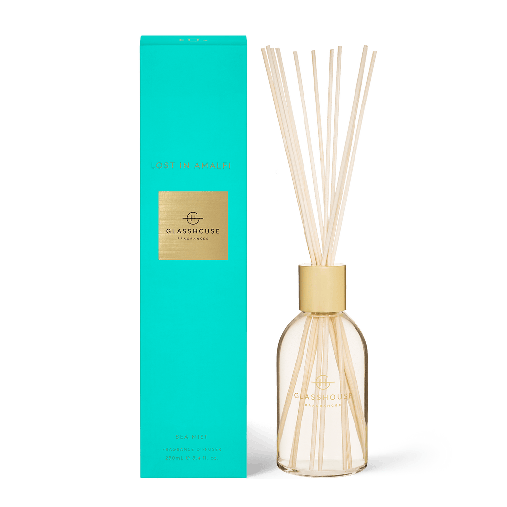 A rectangular turquoise box labeled Lost in Amalfi sits next to a Glasshouse Fragrances diffuser, featuring non-toxic liquid in a clear glass vessel with thin reeds, both displaying the brand logo with gold accents.