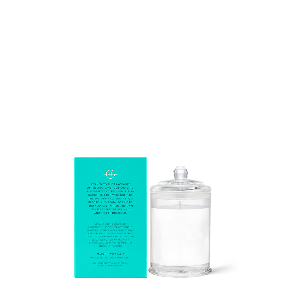 A transparent glass container holds an unused Glasshouse Fragrances - Lost in Amalfi Mini 2.1oz candle, crafted from non-toxic soy blend wax with a ready wick, next to a turquoise box with white text revealing the candles scent and origin.