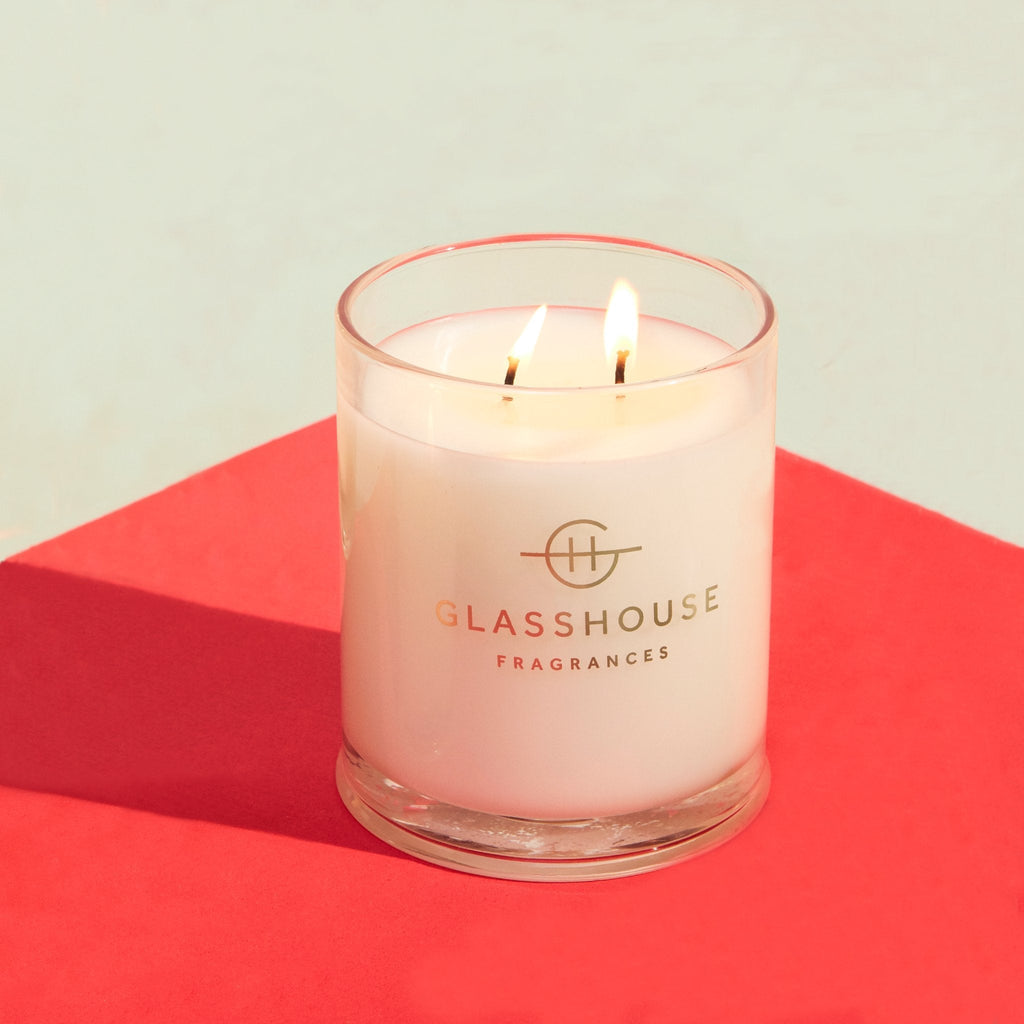 A lit Glasshouse Fragrances - Marseille Memoir 13.4oz Candle, in clear glass with two flames, rests on a red surface against a light background. The gardenia and neroli scent blend perfectly to create an inviting ambiance.