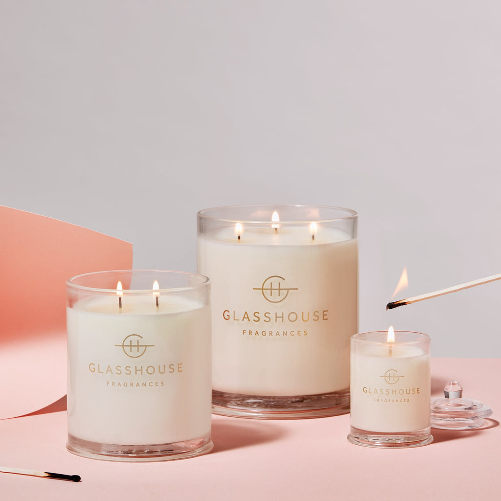 Three Glasshouse Fragrances candles, including the Marseille Memoir mini candle, are on a pink surface, showcasing elegant designs with the brand logo. A lit match is near the mini candle for a neroli fragrance. A piece of pink paper is partially in view behind them.