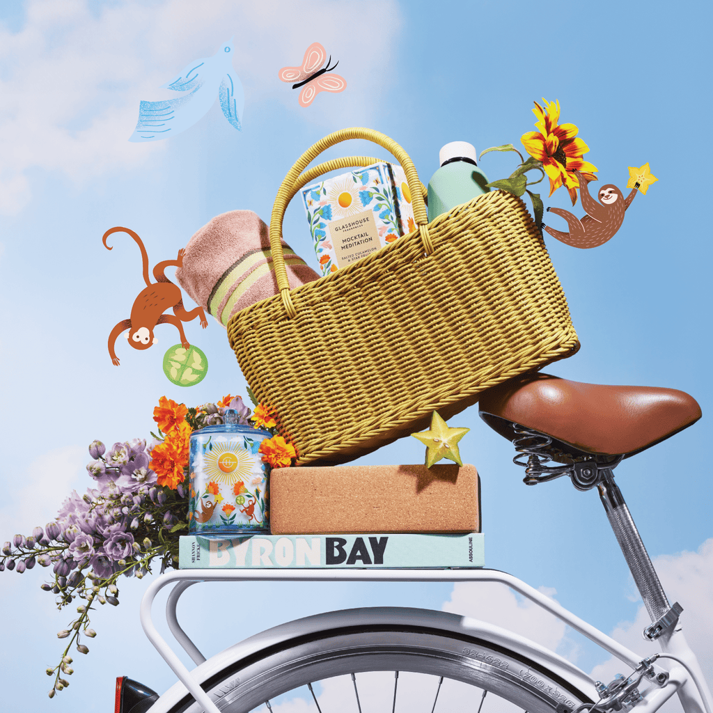A wicker picnic basket with flowers, towels, and drinks sits on a bicycle rack. Books, a can, and a Glasshouse Fragrances - Mocktail Meditation 13.4oz Candle are below, with playful animals and sunflowers against a blue sky backdrop.