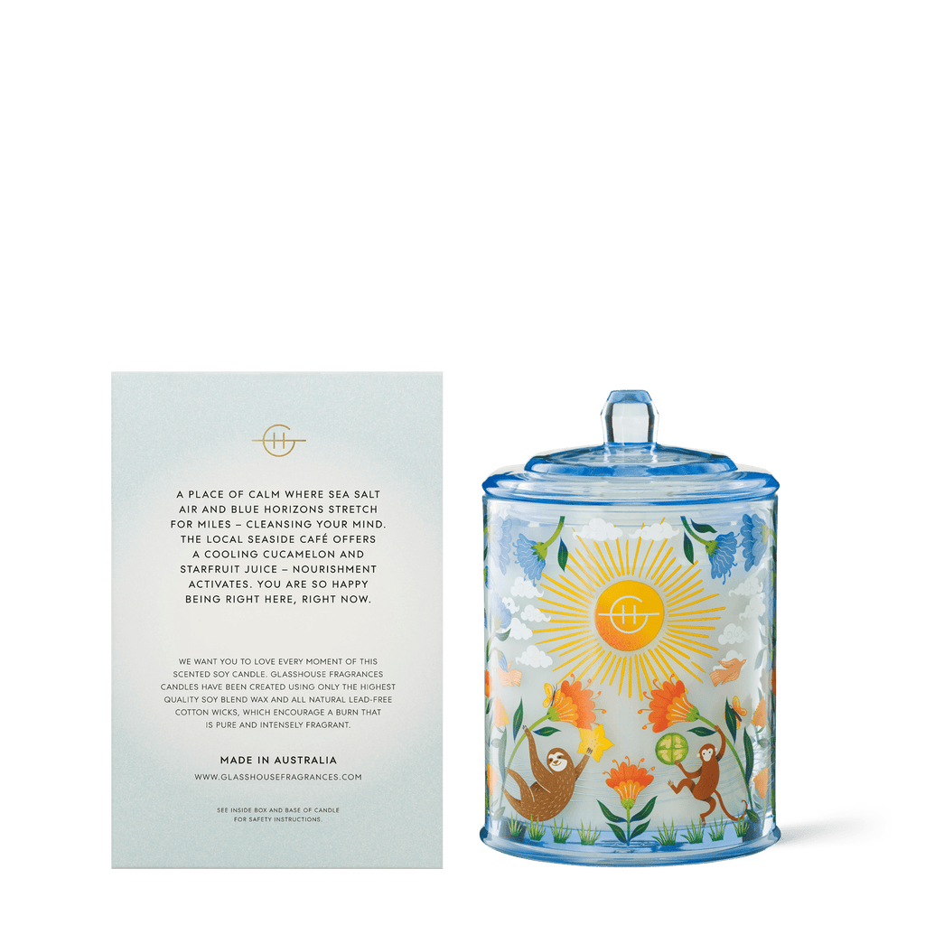 The Glasshouse Fragrances Mocktail Meditation 13.4oz Candle comes in a decorative glass jar with a blue rim and lid, adorned with animals, flowers, and a sun design. A card details its Australian origin and the unique Triple Scented Soy Candle experience.