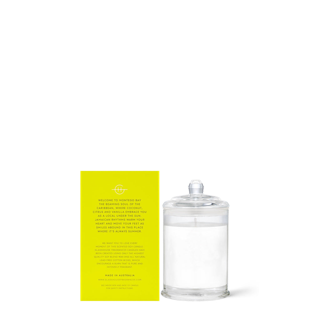 The Glasshouse Fragrances Montego Bay Rhythm Mini 2.1oz Candle, featuring a soy blend wax with coconut & lime fragrance, is displayed beside a bright yellow card with English text. The candle in its clear glass jar with a lid sits elegantly against a plain white background.