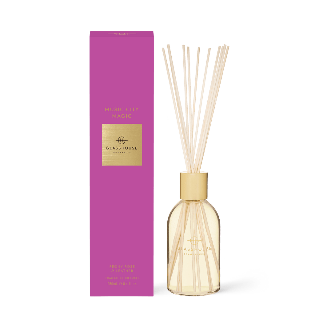 The Glasshouse Fragrances - Music City Magic Diffuser features a transparent bottle with liquid and reed sticks, exuding leather and peony rose notes. It is elegantly paired with a vibrant pink box adorned with gold accents.