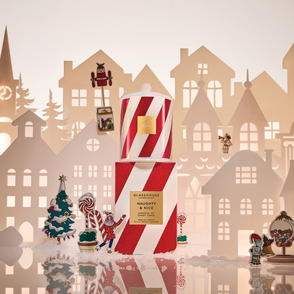 The Glasshouse Fragrances Naughty & Nice Candle, with red and white stripes, sits on its matching box among paper houses, festive holiday décor, a Christmas tree, and miniature figurines.