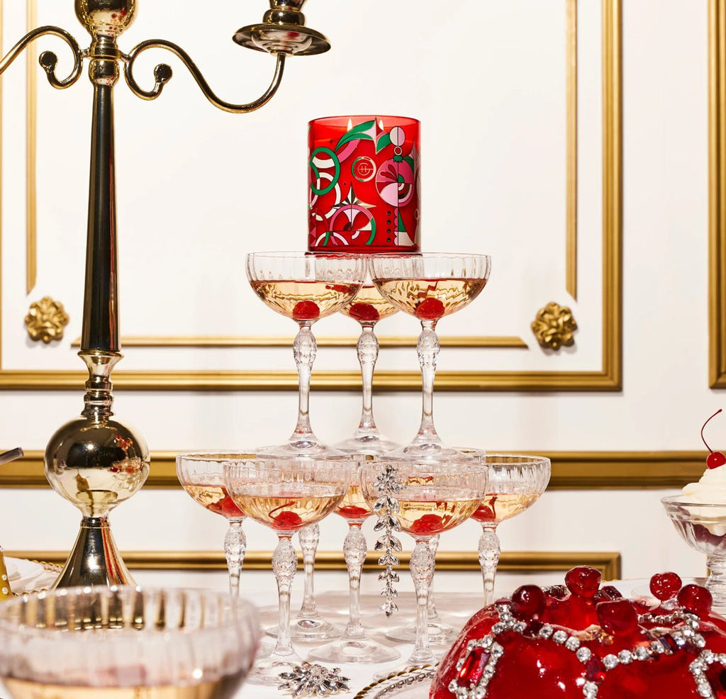 A luxurious table setting features a pyramid of champagne glasses with pink champagne, topped by a Glasshouse Fragrances - Night Before Christmas candle. A silver candelabrum and red berry-topped desserts enhance the charm, filling the air with festive fragrance against an ornate gold-trimmed wall.