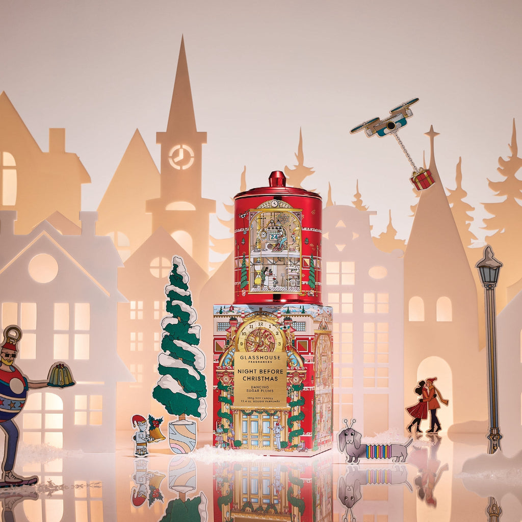 A festive scene displays the Glasshouse Fragrances - Night Before Christmas 13.4oz Candle, set among paper cut-out buildings, a snow-covered tree, holiday figurines, a toy robot, a dog, and a small airplane ornament.