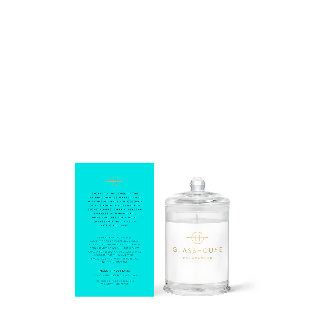 The Glasshouse Fragrances Passion In Positano Mini 2.1oz Candle, in a clear glass container with lid and logo, sits beside a turquoise box with gold text for product and brand info. Its Italian Citrus Bouquet scent refreshes the air.