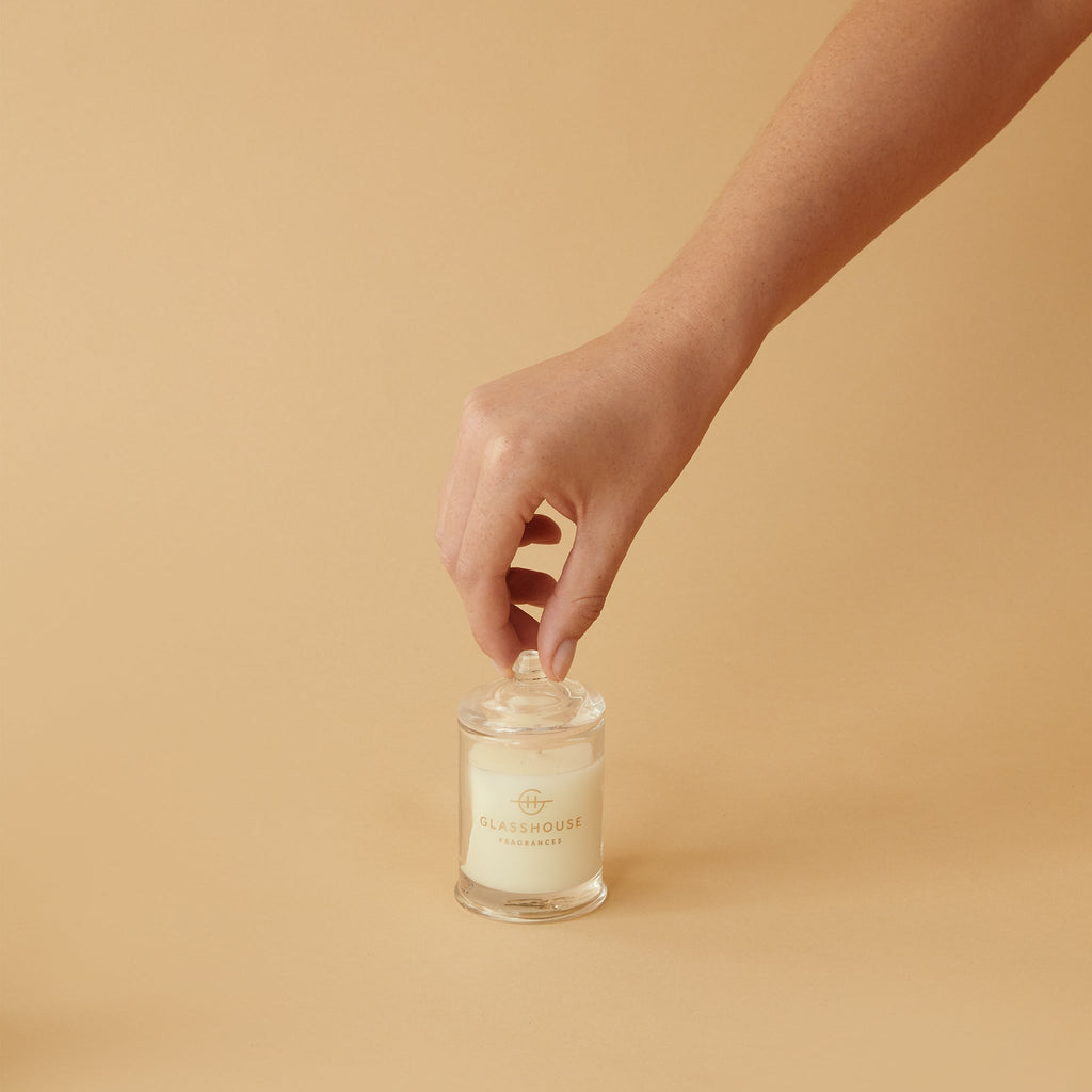 A hand lifts the lid off a Glasshouse Fragrances Rendezvous Mini 2.1oz Candle set against a beige background. The candle, in a clear glass jar with a subtle logo, is crafted from non-toxic soy blend wax for a cleaner burn.