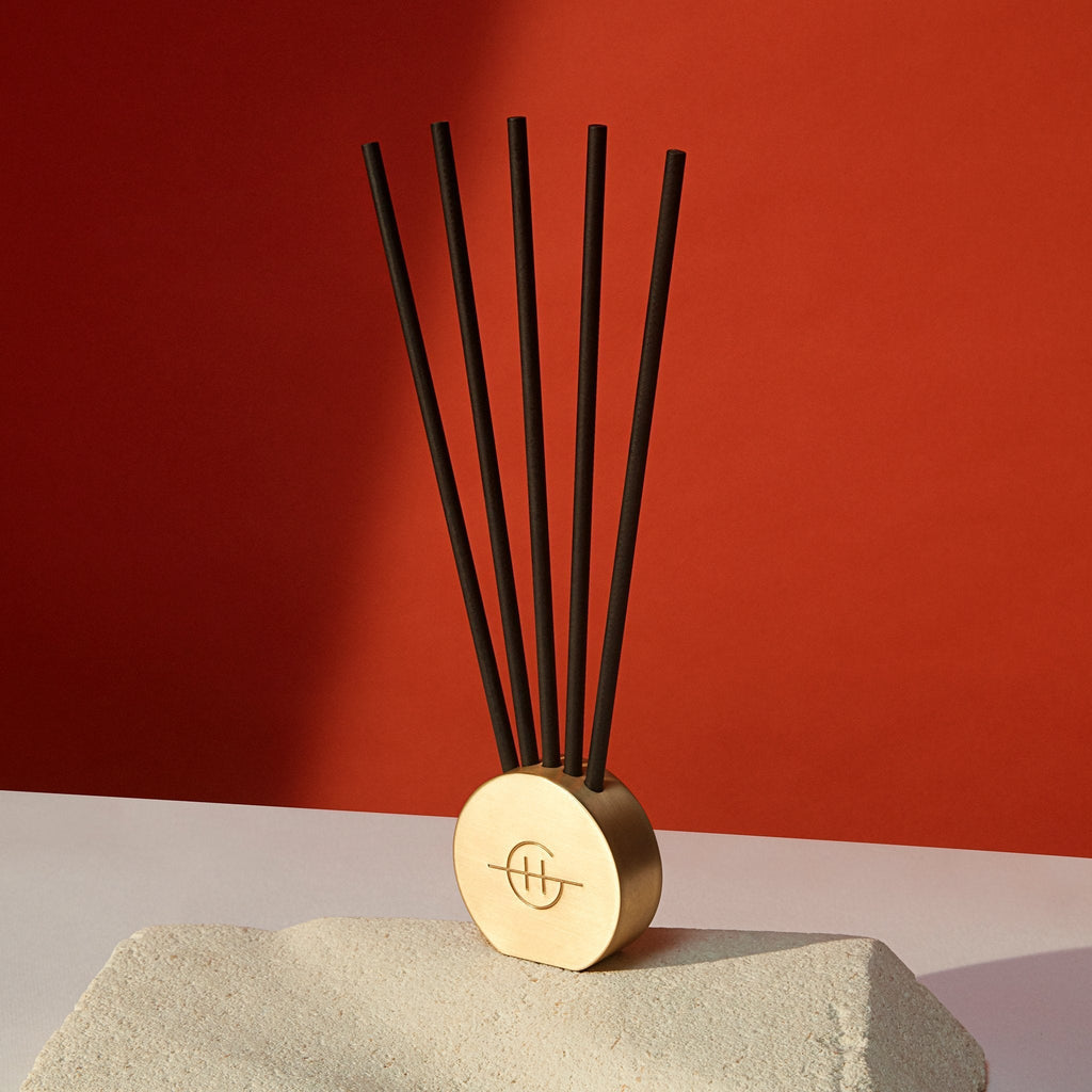 A gold-toned oval base with a decorative symbol holds five vertical black Glasshouse Fragrances - Replacement Scent Stems - Ill Take Manhattan. Enhanced by orchid-scented stems, it rests on a textured beige surface against a vibrant red background.