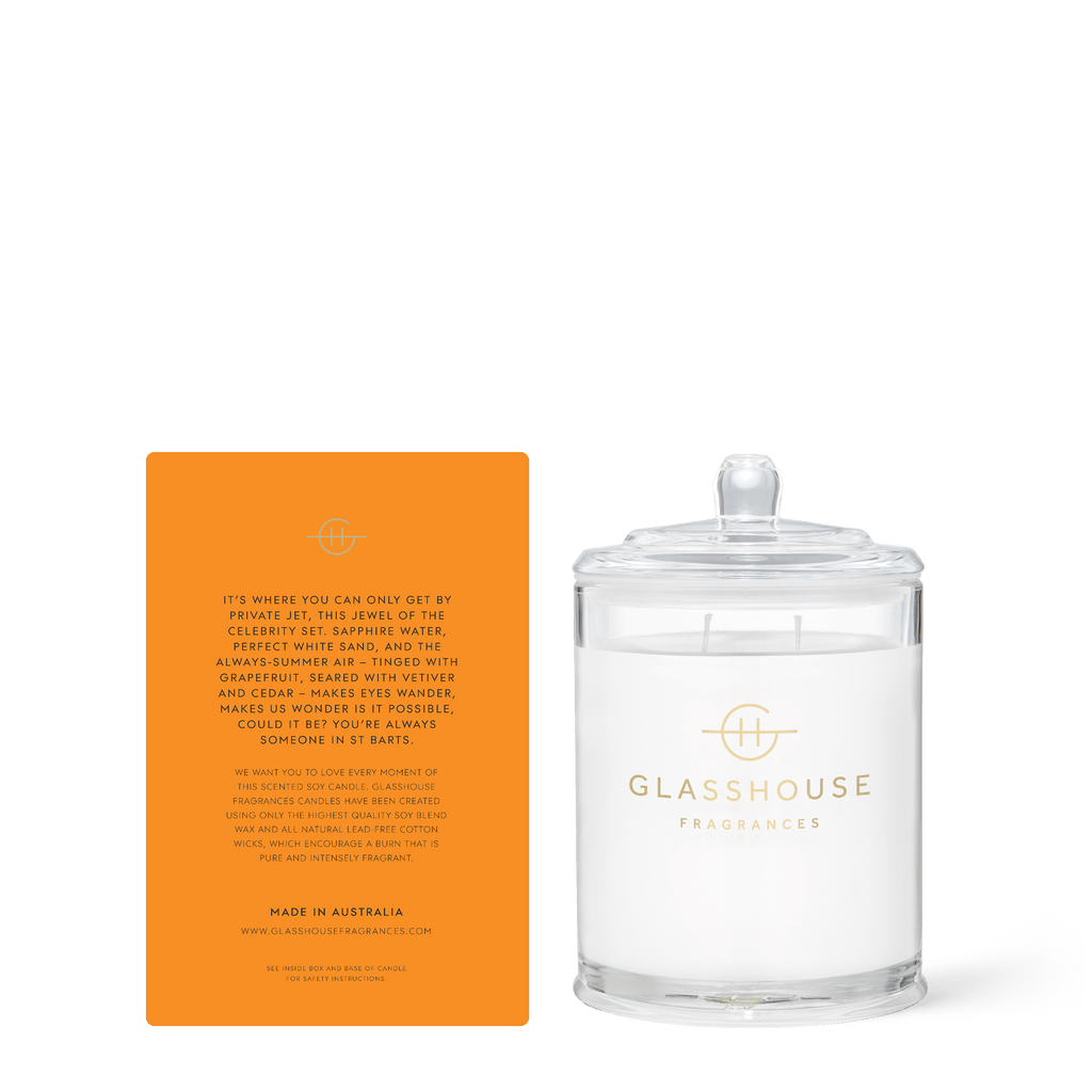 The St Barts Bronze 13.4oz Candle from Glasshouse Fragrances, in clear glass with a branded lid, includes an orange card detailing its Grapefruit scent and the luxurious summer inspiration behind it.