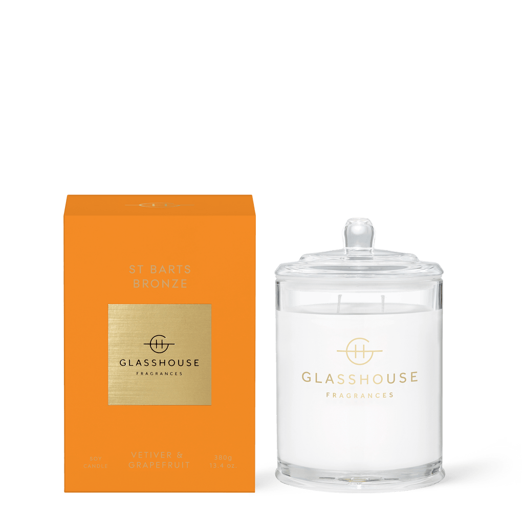 The Glasshouse Fragrances St Barts Bronze Candle (13.4oz) is housed in a sleek orange box with an elegant glass jar and clear lid, featuring a gold label. It fills spaces with the refreshing Vetiver & Grapefruit aroma for sophisticated ambiance.