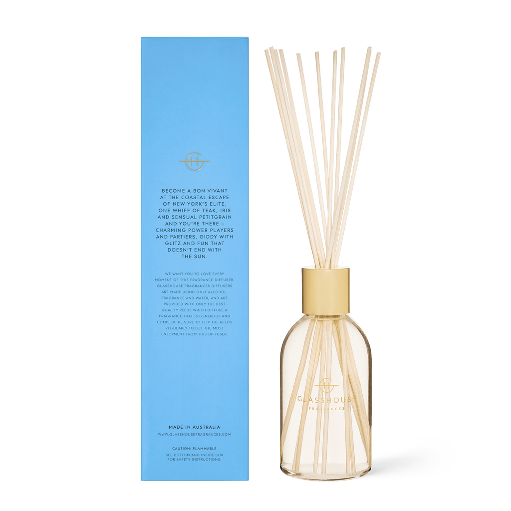 The Glasshouse Fragrances - The Hamptons Diffuser features a glass container with wooden reeds, infused with Teak & Petitgrain. It stands next to a tall blue box with gold logos and text, providing product information, descriptions, and specifications in an elegant design.