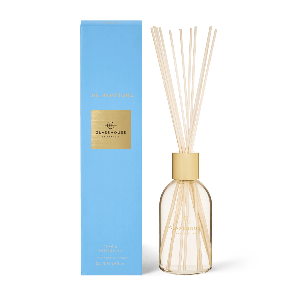 A Glasshouse Fragrances - The Hamptons Diffuser sits beside its blue and gold box, featuring a clear bottle with wooden reeds and gold The Hamptons text. It uses a non-toxic soy blend wax and is enriched with Teak & Petitgrain notes.