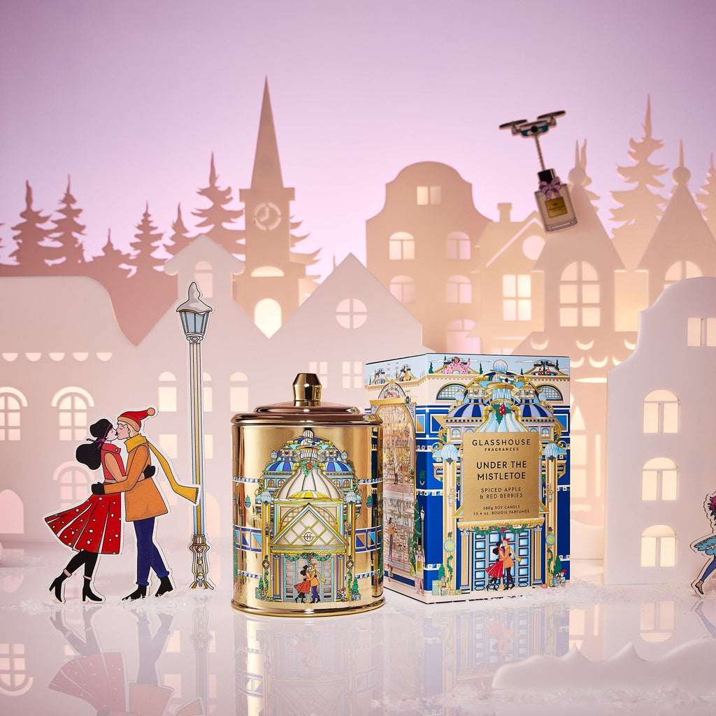 A festive scene featuring the Glasshouse Fragrances - Under the Mistletoe Candle and its box, adorned with holiday art. Behind, a paper cut-out couple kisses by a lamppost amid snowy buildings and trees beneath a pink-purple sky.