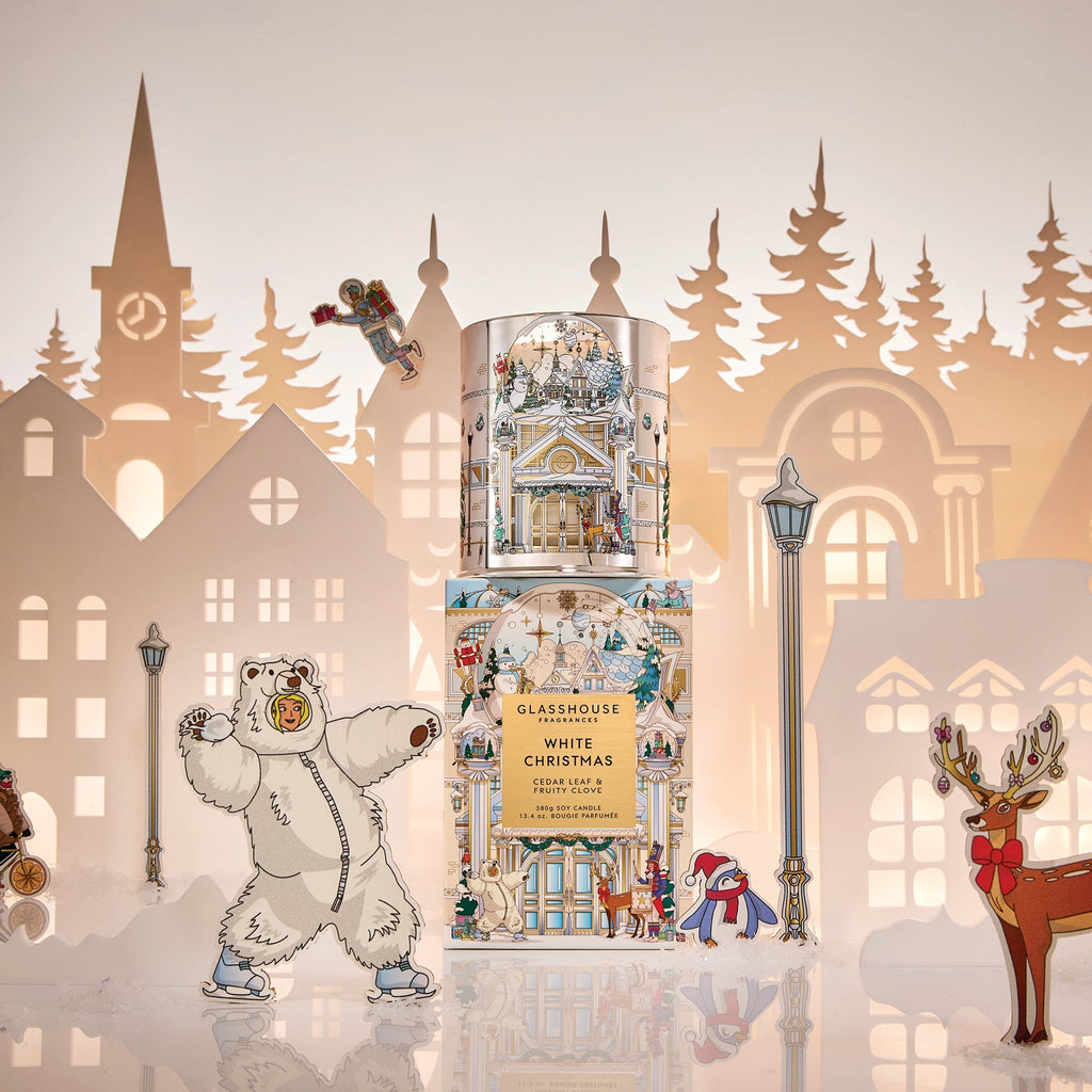 A festive display features the Glasshouse Fragrances - White Christmas 13.4oz Candle at the center, surrounded by paper cutouts of snowy buildings, a dancing polar bear, reindeer, and a penguin in a hat.