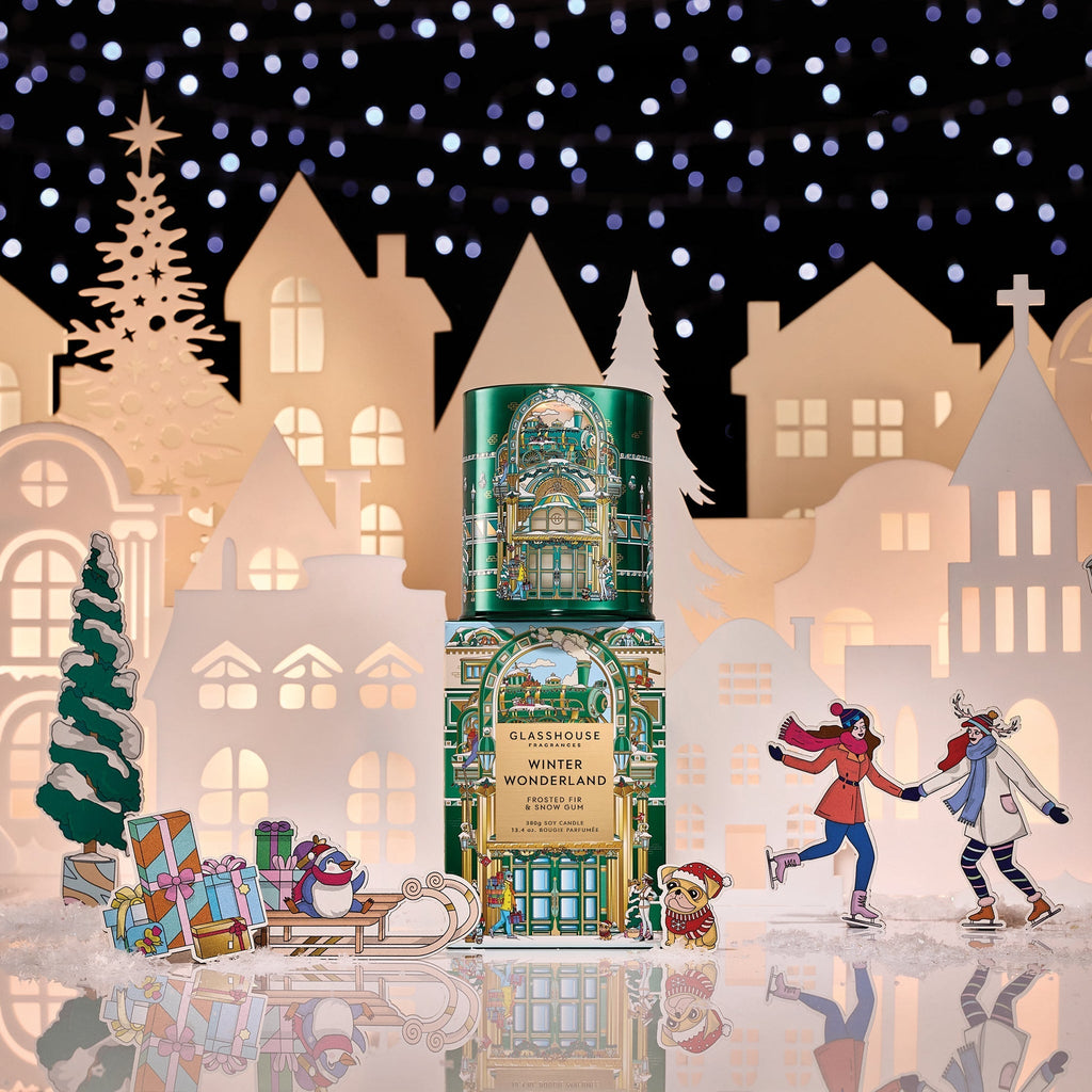 A festive display showcases the Glasshouse Fragrances - Winter Wonderland 13.4oz Candle, surrounded by paper cutouts of snowy houses, a Christmas tree, gifts, and illustrated people ice skating under sparkling lights.