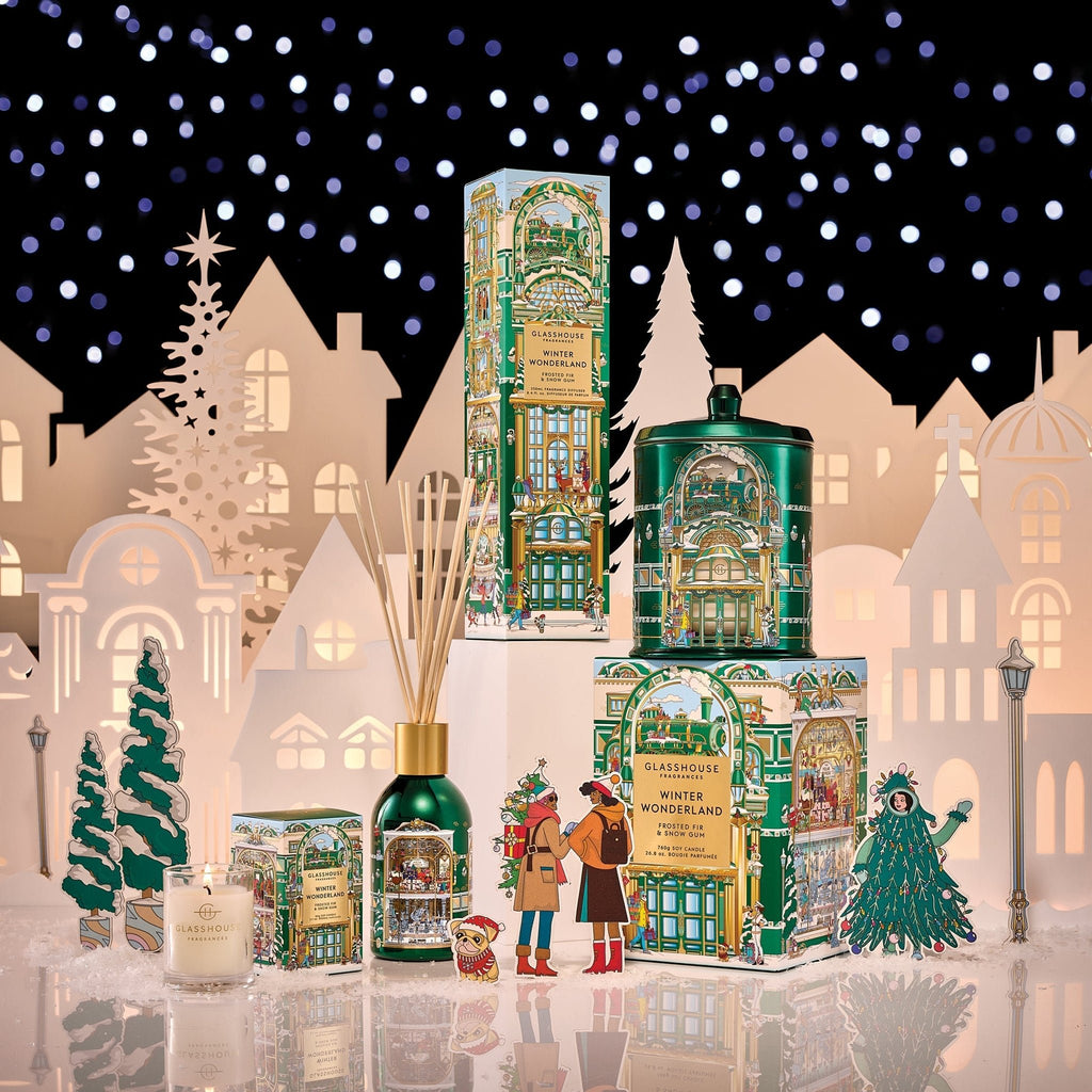 Glasshouse Fragrances' Winter Wonderland 2.1oz Candle is showcased on a snowy surface with a winter village and starry sky backdrop, surrounded by paper trees and festive holiday décor.
