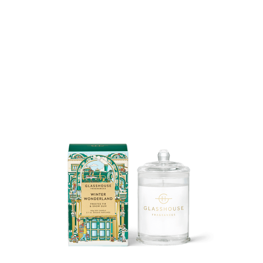 A Glasshouse Fragrances Winter Wonderland 2.1oz candle in a clear jar with a lid is displayed beside its decorative green, gold, and white box featuring a festive building. This triple scented candle is made from a soy blend wax.