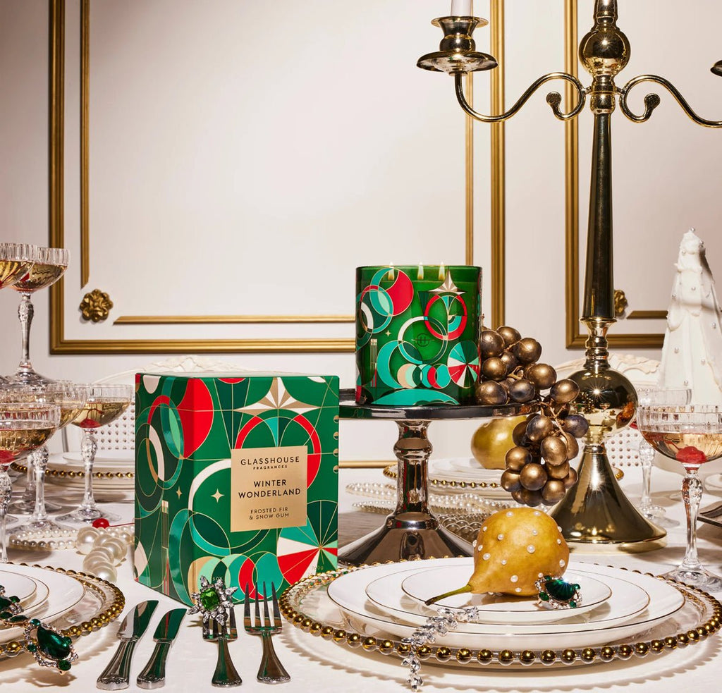 The festive table showcases elegant plates, cutlery, and crystal glasses. A Glasshouse Fragrances Winter Wonderland 26.8oz candle in a decorative box rests on a metal stand. A golden pear and candleholder add luxury with frosted fir hints.