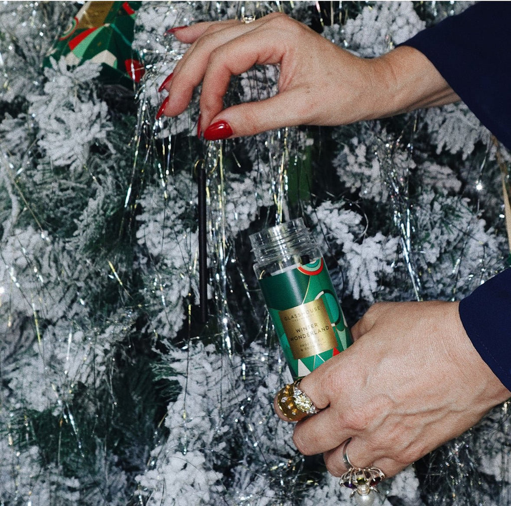 Hands with red nails hang a Glasshouse Fragrances Winter Wonderland Tree Hanging Scent Stem on a snow-covered Christmas tree. The aroma of Fir Balsam fills the air, while the person wearing gold rings adds silver tinsel to the branches, enhancing the festive sparkle.