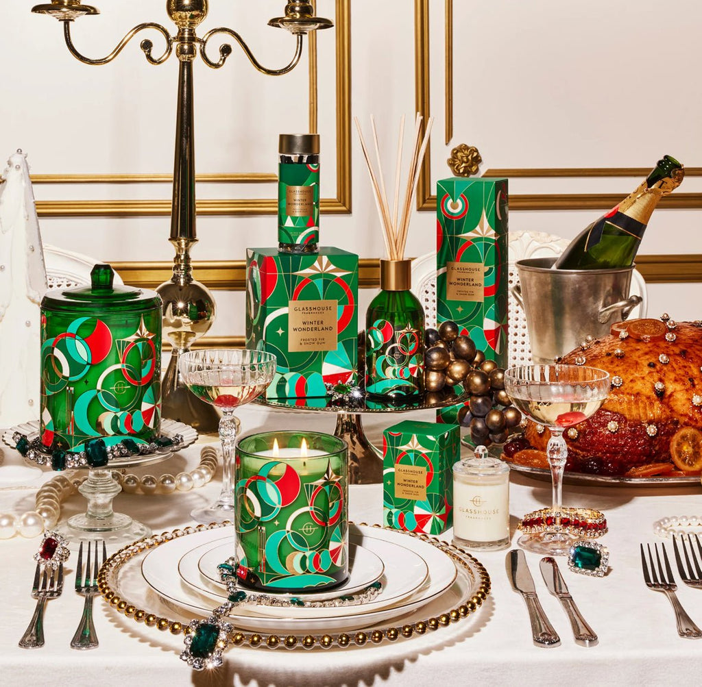 Festive holiday table features green and red luxury gift boxes, a lit candle, champagne on ice, Glasshouse Fragrances Winter Wonderland Tree Hanging Scent Stems for freshness, glasses, decorative beads, a large decorated loaf, and a gold candelabra to complete the elegant setting.