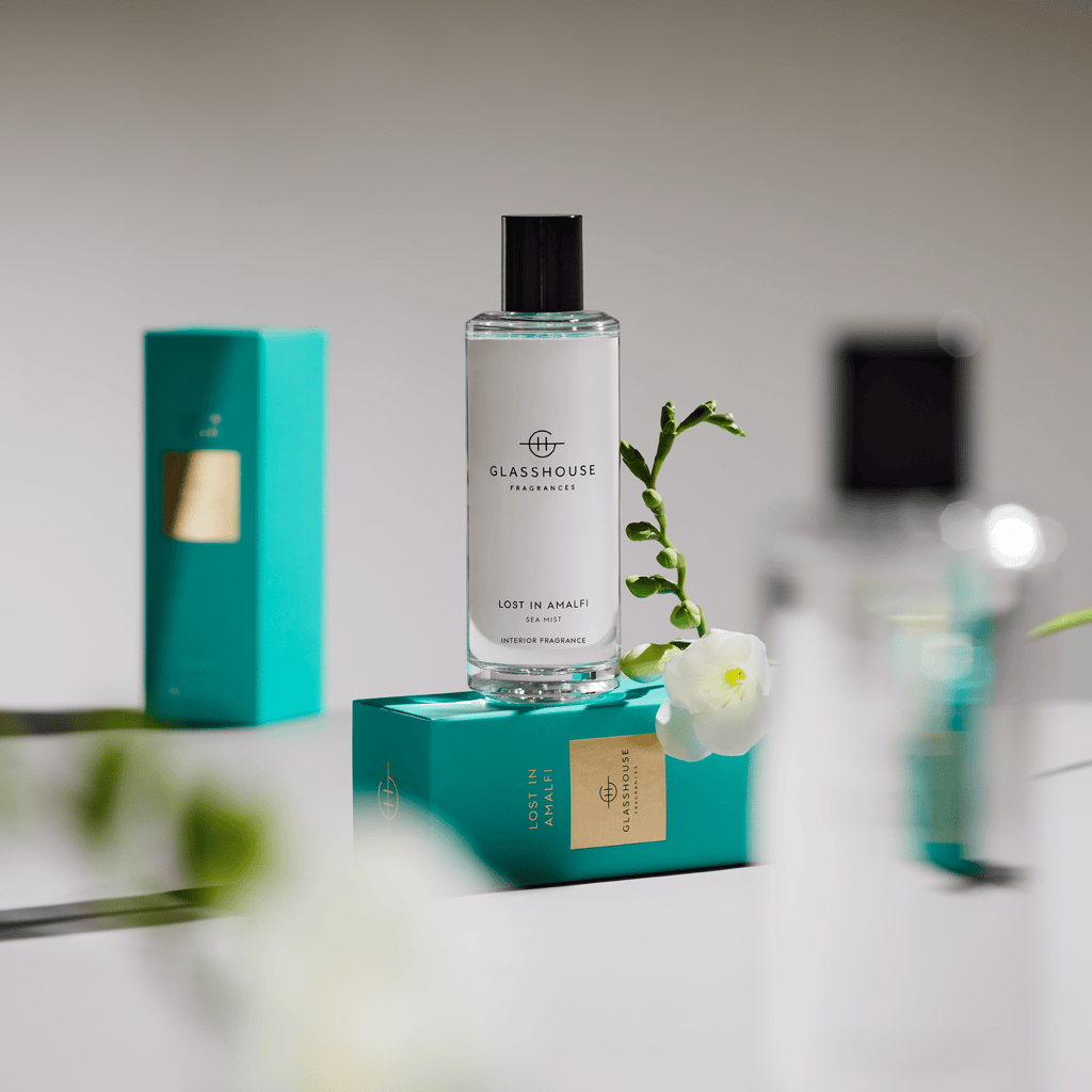 A Glasshouse Fragrances Lost in Amalfi Room Spray bottle rests elegantly on a turquoise box, enhanced by a small white freesia. In the backdrop, another box and a blurred bottle complete the serene ambiance.