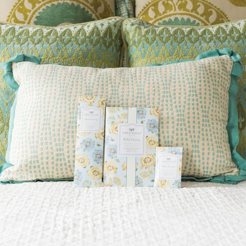 A decorative pillow with a blue and beige pattern rests on a white textured bedspread. In front of the pillow are three Greenleaf Bella Freesia sachets in various sizes, featuring floral designs in blue, yellow, and white.