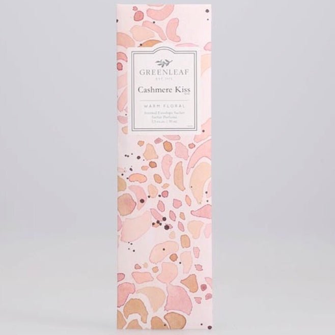 The Greenleaf - Cashmere Kiss Sachet comes in a box with a pastel pink and peach abstract design. The label says Greenleaf Cashmere Kiss, providing a warm, floral fragrance in large sachets to envelop your space elegantly.