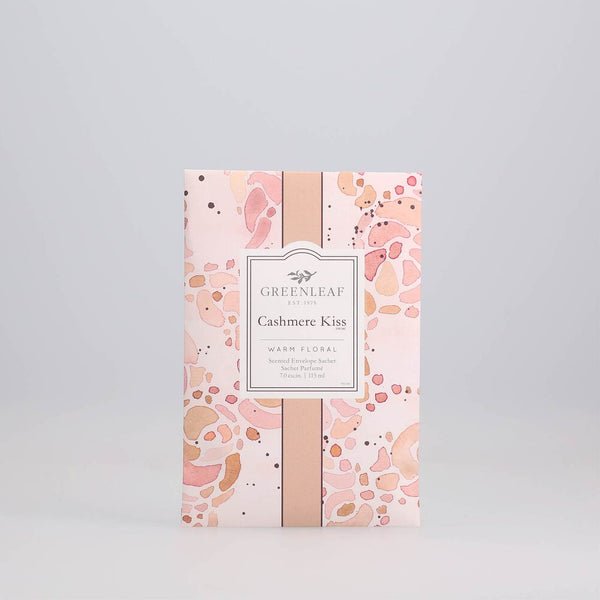 A decorative pink sachet packet by Greenleaf, features abstract peach and beige designs. Labeled Greenleaf - Cashmere Kiss Sachet, it offers a warm cashmere kiss with a delightful floral aroma, elegantly enclosed in a scented envelope.