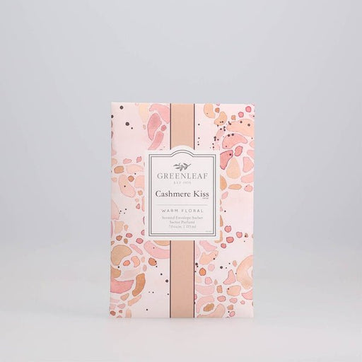 A decorative pink sachet packet by Greenleaf, features abstract peach and beige designs. Labeled Greenleaf - Cashmere Kiss Sachet, it offers a warm cashmere kiss with a delightful floral aroma, elegantly enclosed in a scented envelope.