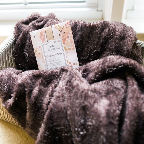 A soft, fluffy brown blanket is draped over a chair, with a Greenleaf Cashmere Kiss Sachet resting on it. The floral fragrance fills the air as sunlight filters through the window with white blinds in the background.