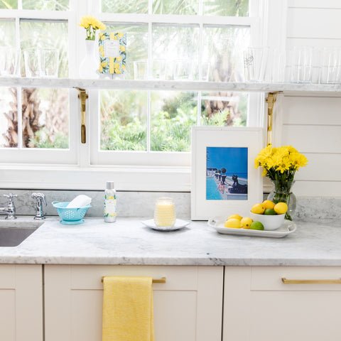 In a bright kitchen, a marble countertop is embellished with a blue colander, candle, and Greenleafs Citron Sol touches like lemons and yellow flowers on a tray. A yellow towel adds warmth to the decor, while glasses are placed on a shelf under a window overlooking the serene garden.