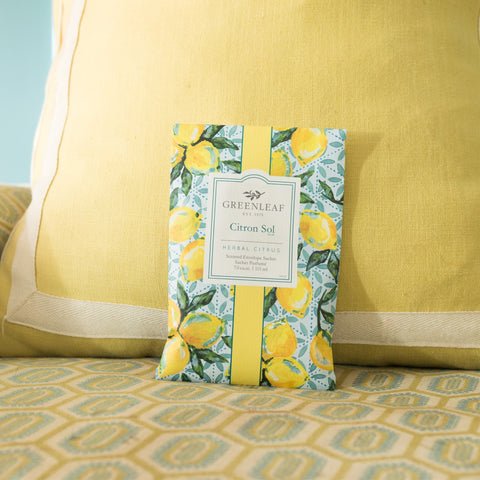 A large sachet, Greenleaf - Citron Sol with a lemon pattern, rests on a yellow geometric cushion. The herbal citrus scent exudes freshness and creates an inviting atmosphere.