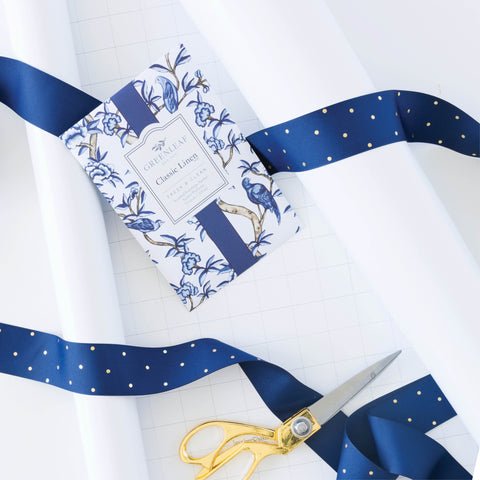 Gift wrapping setup with white paper, gold-handled scissors, and a blue ribbon with gold dots. A booklet titled G. Lalo Classic Linen lies on the paper beside Greenleaf - Classic Linen Sachets, adding scent and elegance to your gift presentation.