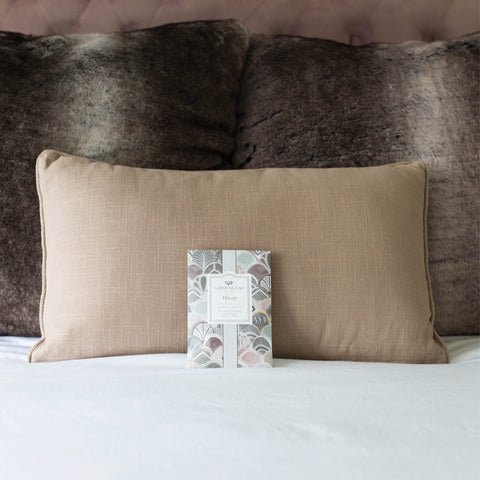 A beige pillow graces the bed amid dark, textured cushions. Nearby sits an elegantly packaged Greenleaf - Haven Sachet, enveloping your space with a soothing fragrance transforming it into a serene haven.